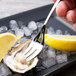 A Chef & Sommelier stainless steel cocktail fork in an oyster shell with a lemon wedge on ice.
