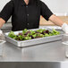 A woman in black gloves places a Hatco stainless steel food pan of lettuce and greens in a countertop warmer.