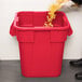 A person pouring food into a red Rubbermaid square trash can.