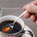 A hand holding an Arcoroc stainless steel demitasse spoon over a cup of coffee.