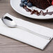 An Arcoroc stainless steel dessert spoon on a white napkin next to a plate of chocolate cake with berries.