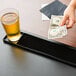 A hand holding a dollar bill on a black bar mat next to a glass of beer.