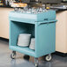 A slate blue Cambro tray and dish cart with plates and utensils on it.