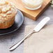 A muffin on a plate next to a Chef & Sommelier stainless steel butter spreader.