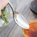 A close up of a Master's Gauge stainless steel teaspoon holding liquid over a jar of orange liquid.