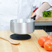 A chef in a white coat uses a Vollrath aluminum sauce pan with a black silicone handle to stir tomatoes over a hot plate.