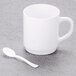 A white mug with a Fineline Tiny Tasters white plastic spoon on it.