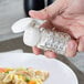 A hand holding a Tablecraft glass shaker with a white cap over a plate.