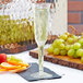 A Fineline clear plastic champagne flute filled with liquid on a table next to a plate of fruit.