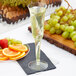 A Fineline clear plastic champagne flute filled with champagne and garnished with an orange slice on a table next to a plate of fruit.