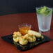 A Fineline black plastic square plate with food next to a glass of clear liquid with lime slices and ice.
