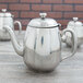 A group of three Vollrath stainless steel teapots on a wood surface.