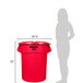 A woman standing next to a red Rubbermaid BRUTE trash can with a red lid.