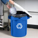 A person putting a plastic container with white lid into a blue Rubbermaid recycling can.