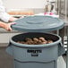 A woman opening a Rubbermaid BRUTE 55 gallon grey trash can with a grey lid to reveal potatoes.