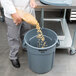 A man in a chef's uniform pouring pasta into a gray Rubbermaid trash can with a lid.