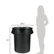 A woman standing next to a black Rubbermaid trash can with a black lid.