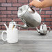 A person pouring coffee into a stainless steel teapot.
