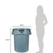 A woman standing next to a large grey Rubbermaid trash can with a lid.