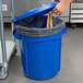 A hand pulling a plastic bag into a blue Rubbermaid BRUTE trash can.
