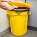 A hand reaching out to open a Rubbermaid yellow trash can lid.