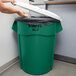 A woman putting a black bag into a green Rubbermaid BRUTE trash can with a white lid in a corporate office cafeteria.