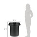A woman standing next to a black Rubbermaid BRUTE trash can with a black lid.
