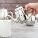 A hand pouring coffee from a Vollrath stainless steel teapot into a cup.