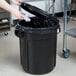 A person's hand putting foil in a Rubbermaid black round trash can with a black lid.