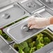 A gloved hand using a Choice stainless steel pan cover to take broccoli out of a tray.