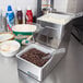A San Jamar stainless steel condiment bar on a counter with bowls of food and a metal container of chocolate chips.