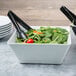 A white melamine square bowl filled with salad with black tongs in it.