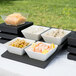 A Vollrath Cubic wood display stand with four black bowls filled with food on a table outdoors.