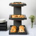 A Vollrath three tiered wood stand with bread and pastries on a table.