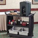 A black Carlisle plastic utility cart with trays holding a beverage dispenser and cookies.