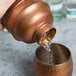 A person using an American Metalcraft antique copper cobbler cocktail shaker to pour water into a cup.