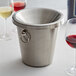A silver Franmara wine tasting spittoon with a ring on top on a table next to wine glasses.