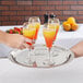 A hand holding a Vollrath stainless steel serving tray with glasses of orange and yellow liquid.