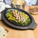 A Lodge pre-seasoned cast iron fajita skillet with grilled chicken, peppers, and onions on it with a wood underliner.