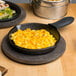 A Lodge mini cast iron skillet of macaroni and cheese on a wooden table with a black silicone handle holder.