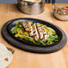 A plate of grilled chicken, peppers, and tortillas on a Lodge oval cast iron fajita skillet with a wood underliner.