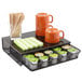 A black Safco coffee pod organizer with orange coffee cups and green and white striped napkins on it.