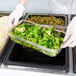 A gloved hand holding a Cambro plastic food container filled with broccoli.