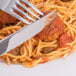 A Oneida Scroll stainless steel dinner knife and fork cutting into spaghetti with sauce.