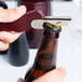 A person using a Franmara Boomerang Waiter's Corkscrew with a burgundy handle to open a brown bottle of beer.