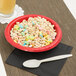 A bowl of cereal with a spoon and a glass of juice on a table with a Classic Red paper bowl filled with cereal.