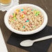 A white paper bowl filled with colorful cereal on a table with a white plastic spoon.