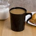 A black Fineline Flairware plastic mug filled with brown liquid on a table with pastries.