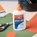 A white bottle of Elmer's Glue-All on a table with a group of colored paper and a pair of scissors.