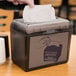 A black and grey Just1 Mini Interfold Tabletop Napkin Dispenser with white napkins in it.
